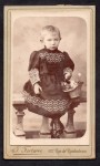 Portret dziewczynki z krótkimi włosami (czarna, haftowana sukienka, koszyczek) Fotografia na kartoniku firmowym Paris [ca. 1885]