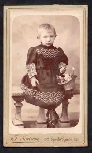 Portret dziewczynki z krótkimi włosami (czarna, haftowana sukienka, koszyczek) Fotografia na kartoniku firmowym Paris [ca. 1885]