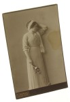 Portret młodej kobiety, stojący, przepaska na głowie, kwiaty w dłoni Fotografia na tekturce firmowej / albumin (Poznań ca 1900 K. Ignatowicz)