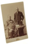 Portret dwójki dojrzałych mężczyzn, jeden siedzi, książki na stole, książka w dłoni Fotografia na tekturce firmowej / albumin (Wiedeń ca 1900 Adele)
