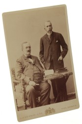Portret dwójki dojrzałych mężczyzn, jeden siedzi, książki na stole, książka w dłoni Fotografia na tekturce firmowej / albumin (Wiedeń ca 1900 Adele)