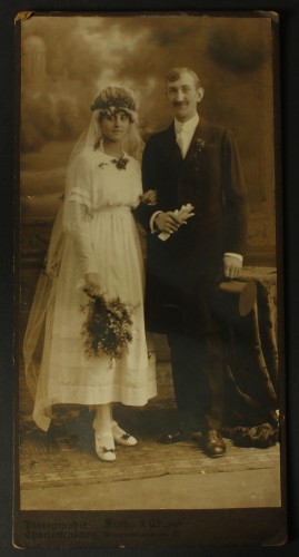Portret młodej pary, stojący, cylinder, rękawiczki, kwiaty Fotografia na tekturce firmowej / odbitka srebrowożelatynowa (Charlottenburg ca 1900 Atelier Fuchs and Co.)