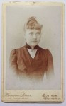 Portret młodej kobiety - popiersie, brosza, włosy upięte, grzywka Fotografia na kartoniku firmowym ca. 1900 / Niemcy