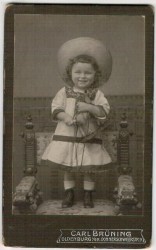 Portret dziewczynki w kapeluszu - stojący, konik - zabawka w rękach Fotografia na kartoniku firmowym (atelier Carla Bruninga Oldenburg) [ca 1910]