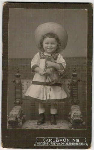 Portret dziewczynki w kapeluszu - stojący, konik - zabawka w rękach Fotografia na kartoniku firmowym (atelier Carla Bruninga Oldenburg) [ca 1910]